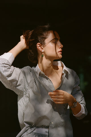 Woman in a Connecticut Crest Striped Shirt - Forest adjusts her hair, standing in sunlight against a dark background.