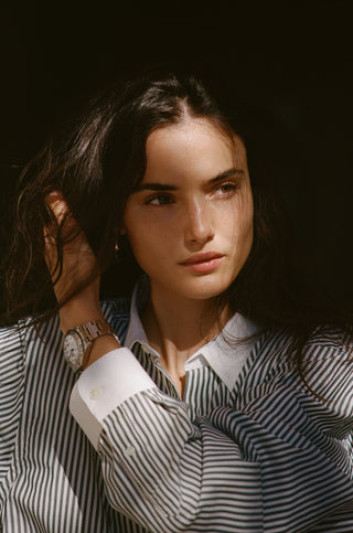 Woman with long dark hair wearing a Connecticut Crest Striped Shirt - Forest, looking to the side, hand in her hair, and wearing a watch.