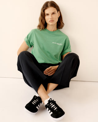 A woman wearing the Disco T-Shirt - Kelly, made from 100% cotton, and black pants sits on the floor against a beige wall, looking at the camera.