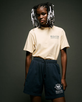 Young woman with beaded braids wears the NY Racquet Club T-Shirt in Almond/Navy, made from 100% USA-grown cotton, paired with navy shorts, standing against a dark background.