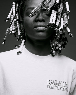 Black and white photo of a person with beaded braids, wearing the NY Racquet Club T-Shirt - Almond/Navy, made from 100% USA grown cotton.