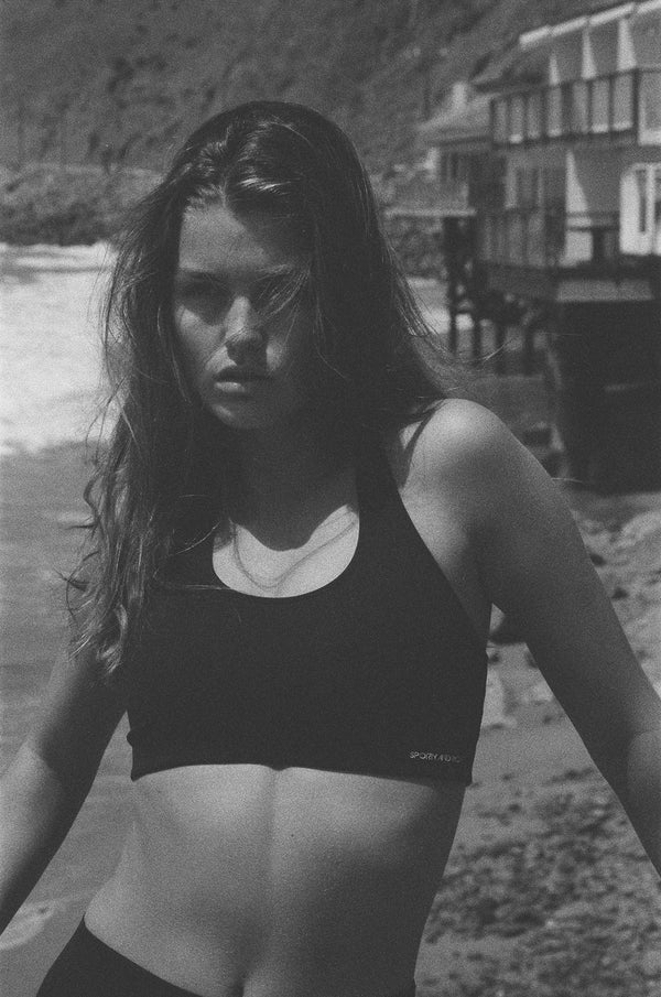 Wearing the Sports Bra N.03 - Navy, a woman enjoys comfort and support as she stands on the beach with waves and buildings in the background.