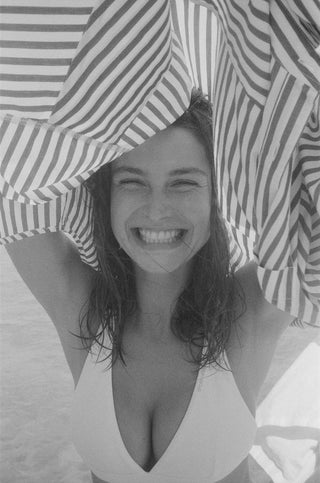 A woman in a bikini smiles brightly outdoors, holding the SRC Oversized Shirt - Chocolate Striped above her head like an oversized shirt.