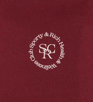 The SRHWC Track Shorts in Merlot is crafted from a red textured, high-quality fabric featuring a white circular logo that reads Sporty & Rich Health & Wellness Club SRC. These shorts boast an elastic waistband for optimal comfort and style, making them the perfect match for various outfits.
